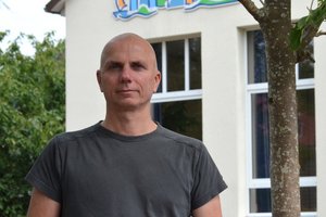 Ein Mann ohne Haare steht neben einem Baum vor einem weißen Haus