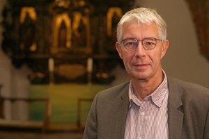 Ein weißhaariger Mann mit Brille schaut in die Kamera, im Hintergrund der Altarraum einer Kirche